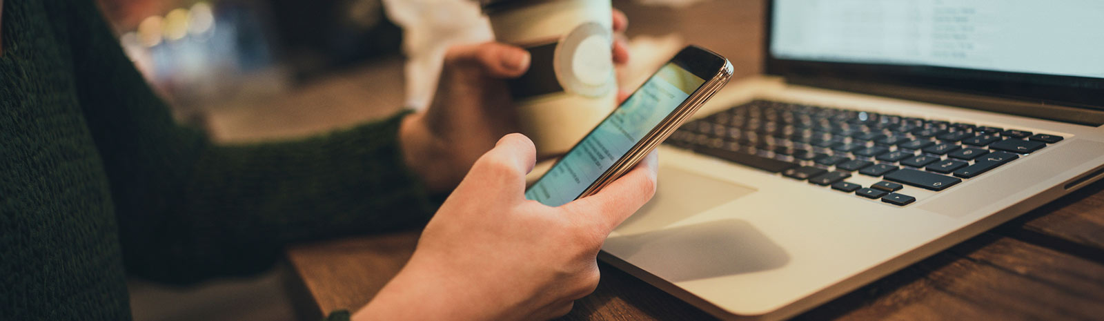 Person holding phone and coffee sitting in front of laptop.