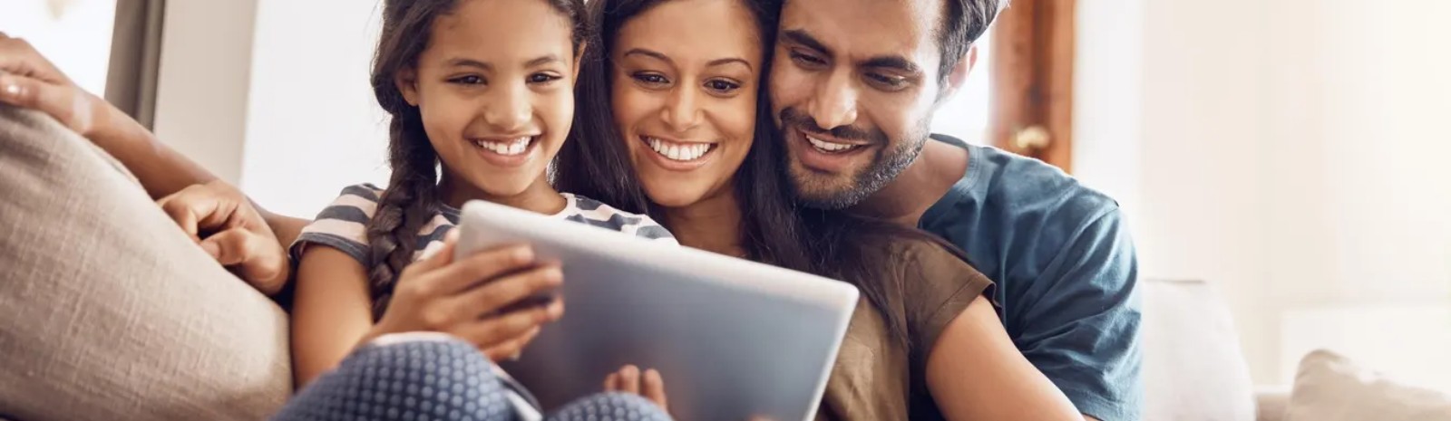 a family of three sitting on a couch looking at a tablet.